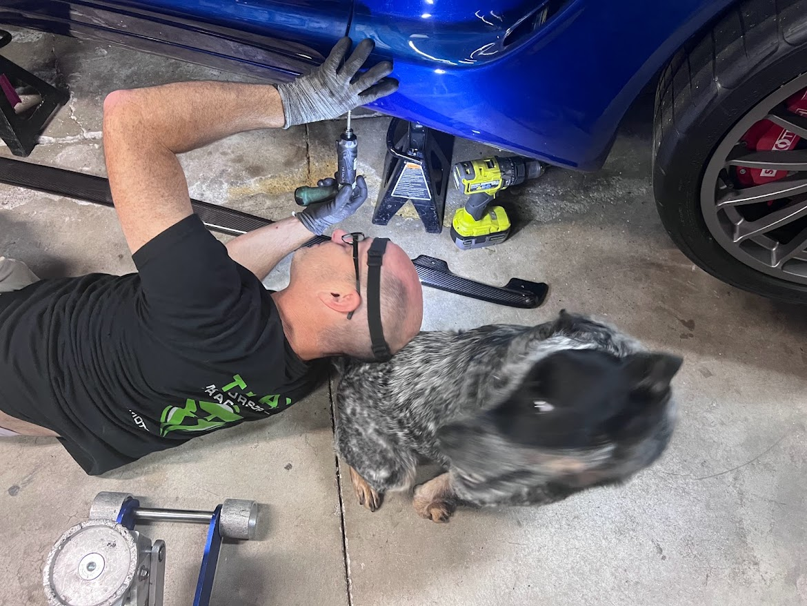 Mechanic working under a blue Corvette with the shop dog nearby
